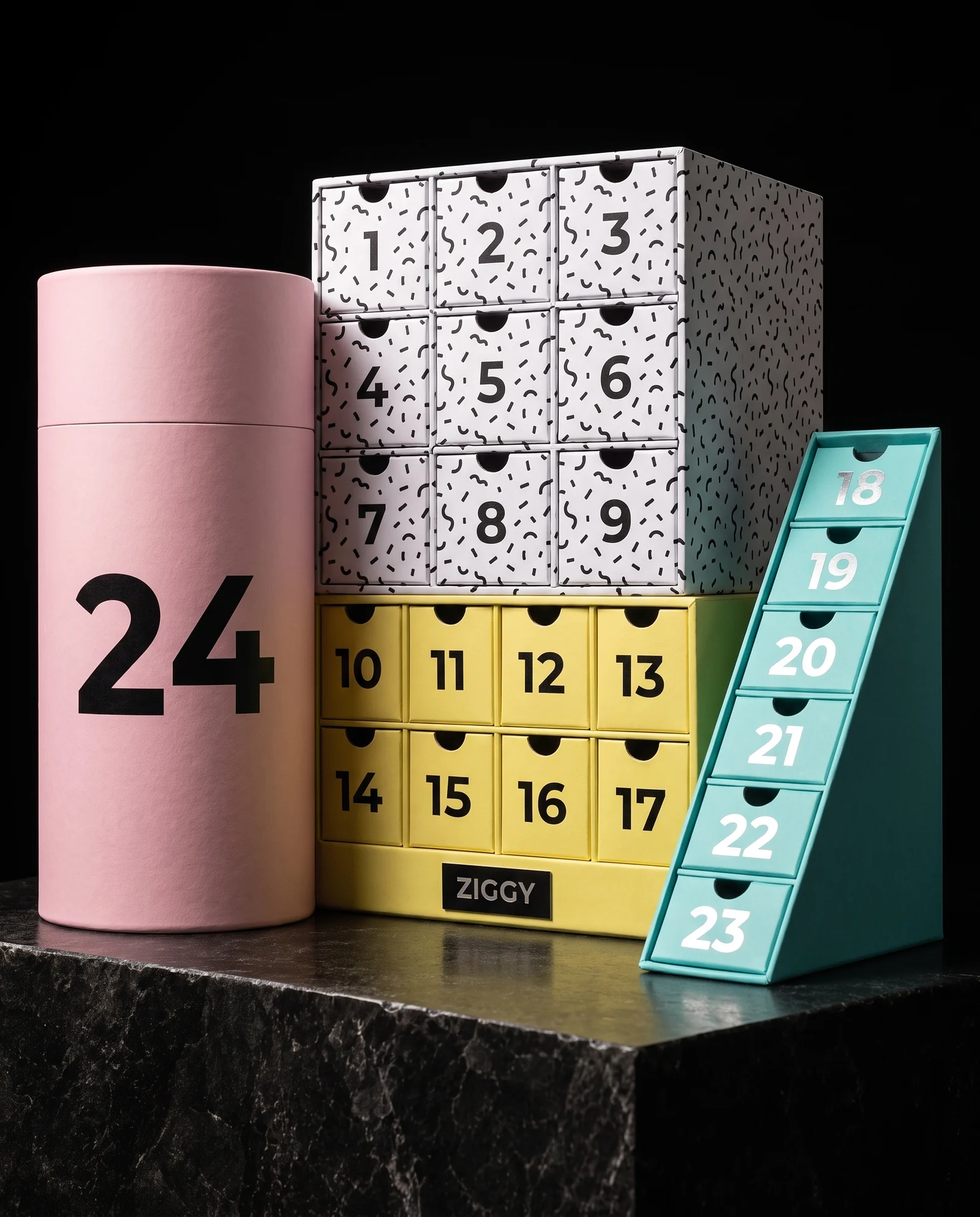 A playful Memphis-style advent calendar: a pink tube marked 24, a black-and-white squiggle cube with drawers 1 to 9, a yellow block with drawers 10 to 17 and a turquoise wedge with drawers 18 to 23, plus a small black ZIGGY plate.