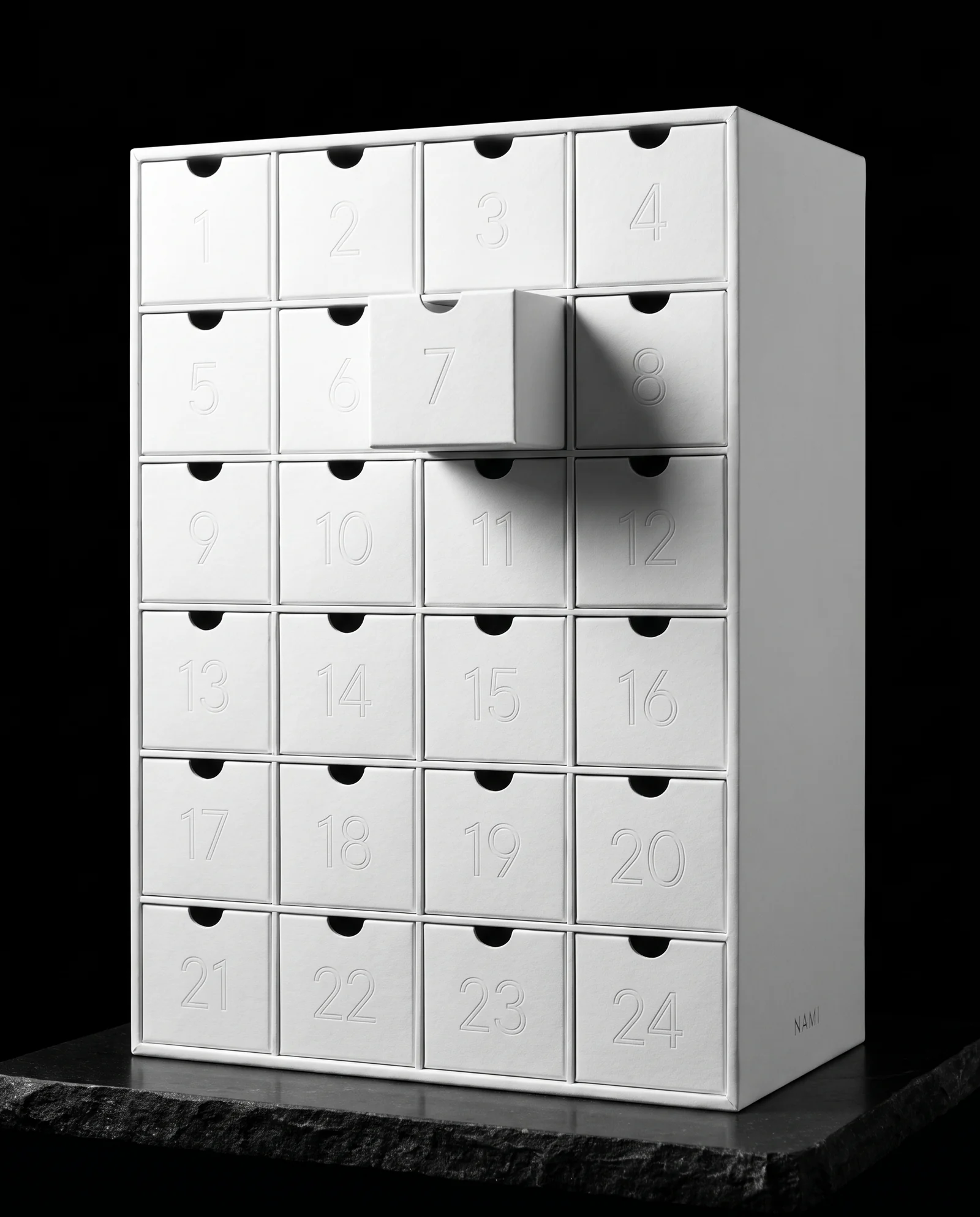 A tall white advent calendar with 24 flush drawers whose numerals are blind debossed with no ink, drawer 7 pulled slightly open, and a tiny matte silver word NAMI on the side.