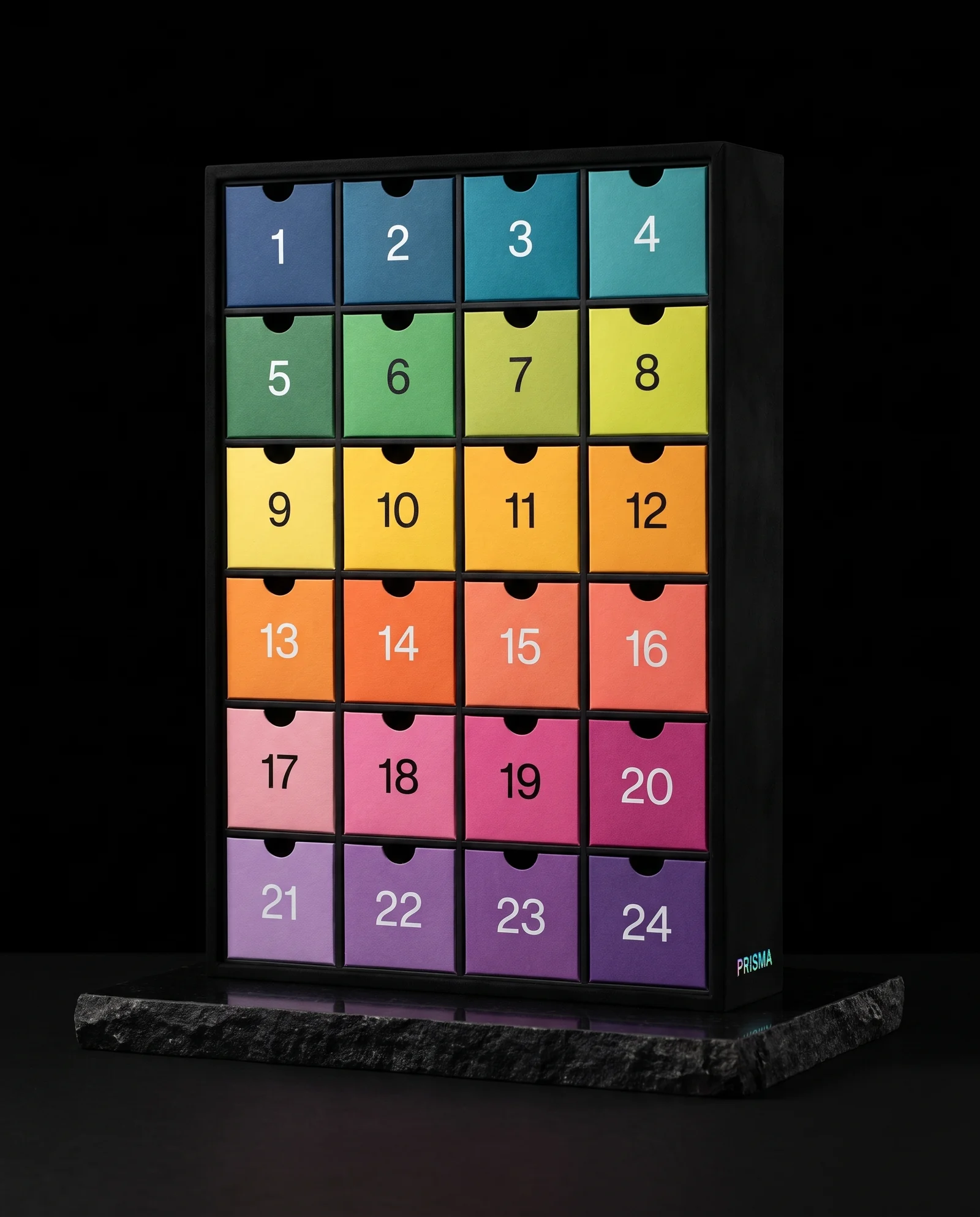 A slim black soft-touch advent calendar frame holding 24 drawers in a stepped gradient of coloured papers from blue through green, yellow, orange, coral and pink to violet, with white and black numerals and a small holographic PRISMA wordmark.