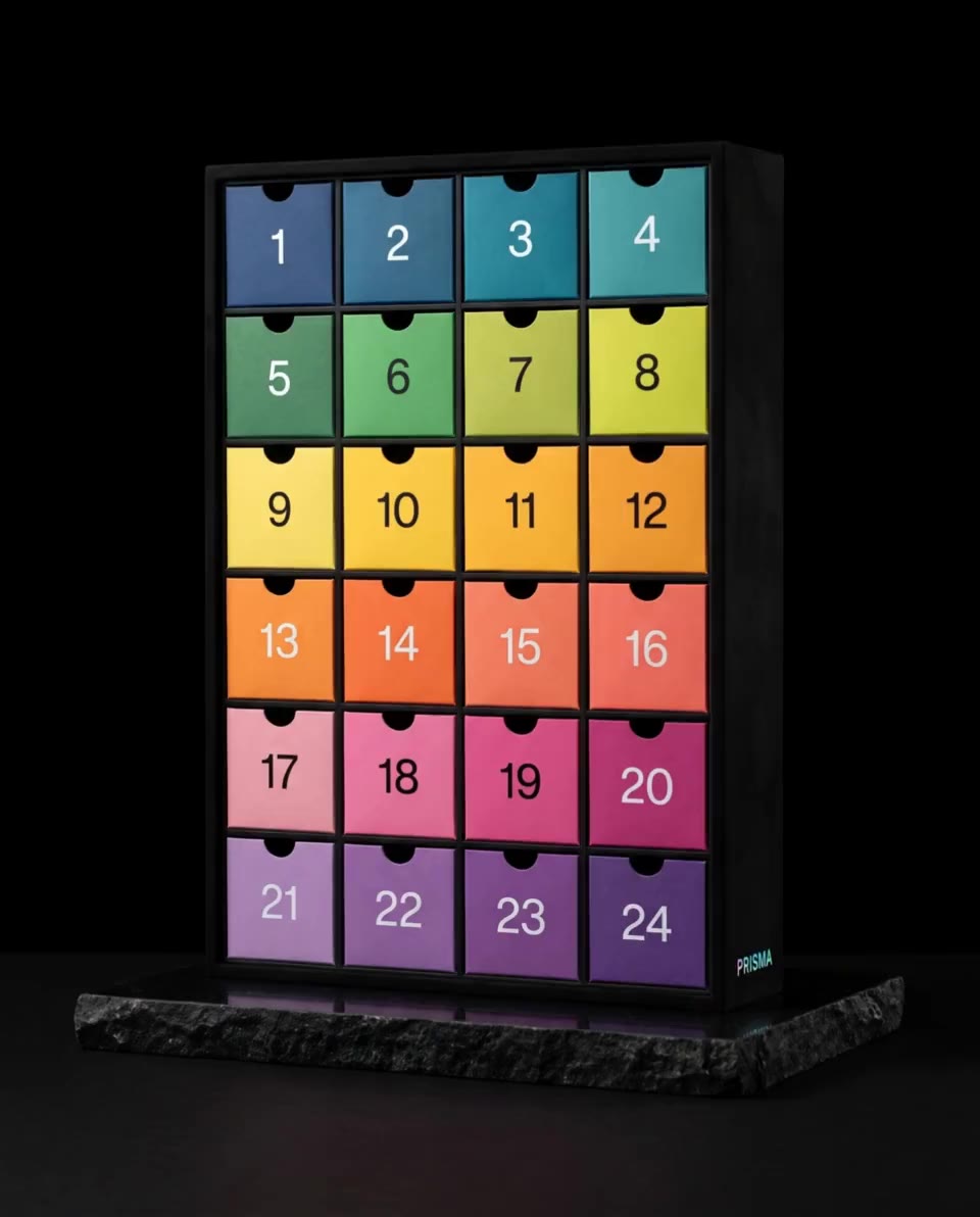 A slim black advent calendar with 24 drawers in a stepped rainbow of coloured papers; the violet drawer 24 slides open a few centimetres and closes again.