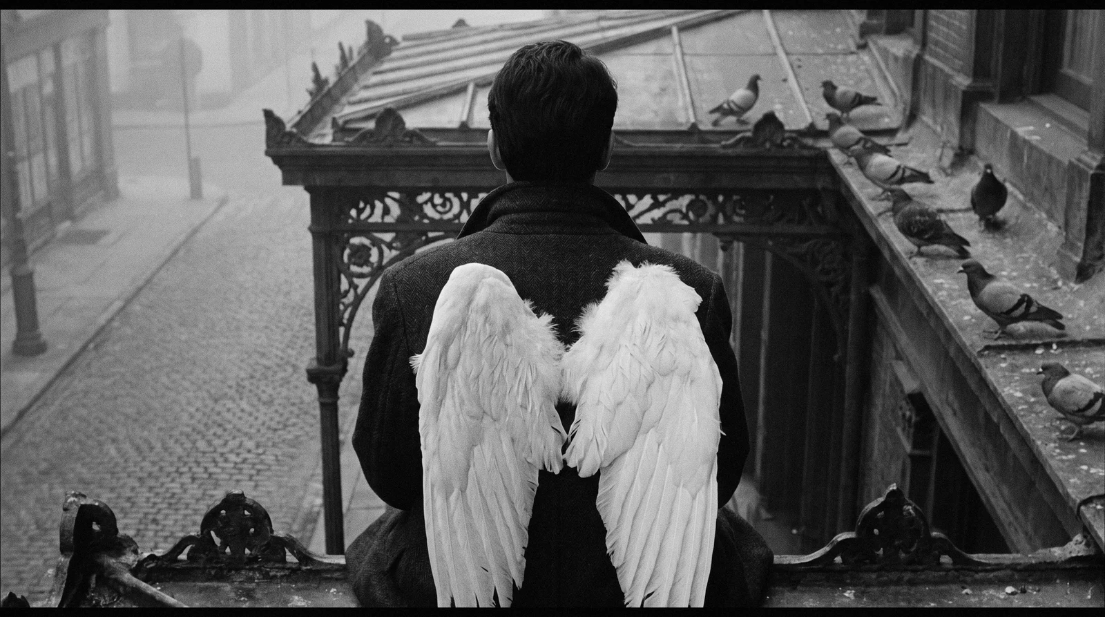 A figure with white wings seen from behind, sitting on a cast-iron cornice above a cobbled street, pigeons beside him, in black and white