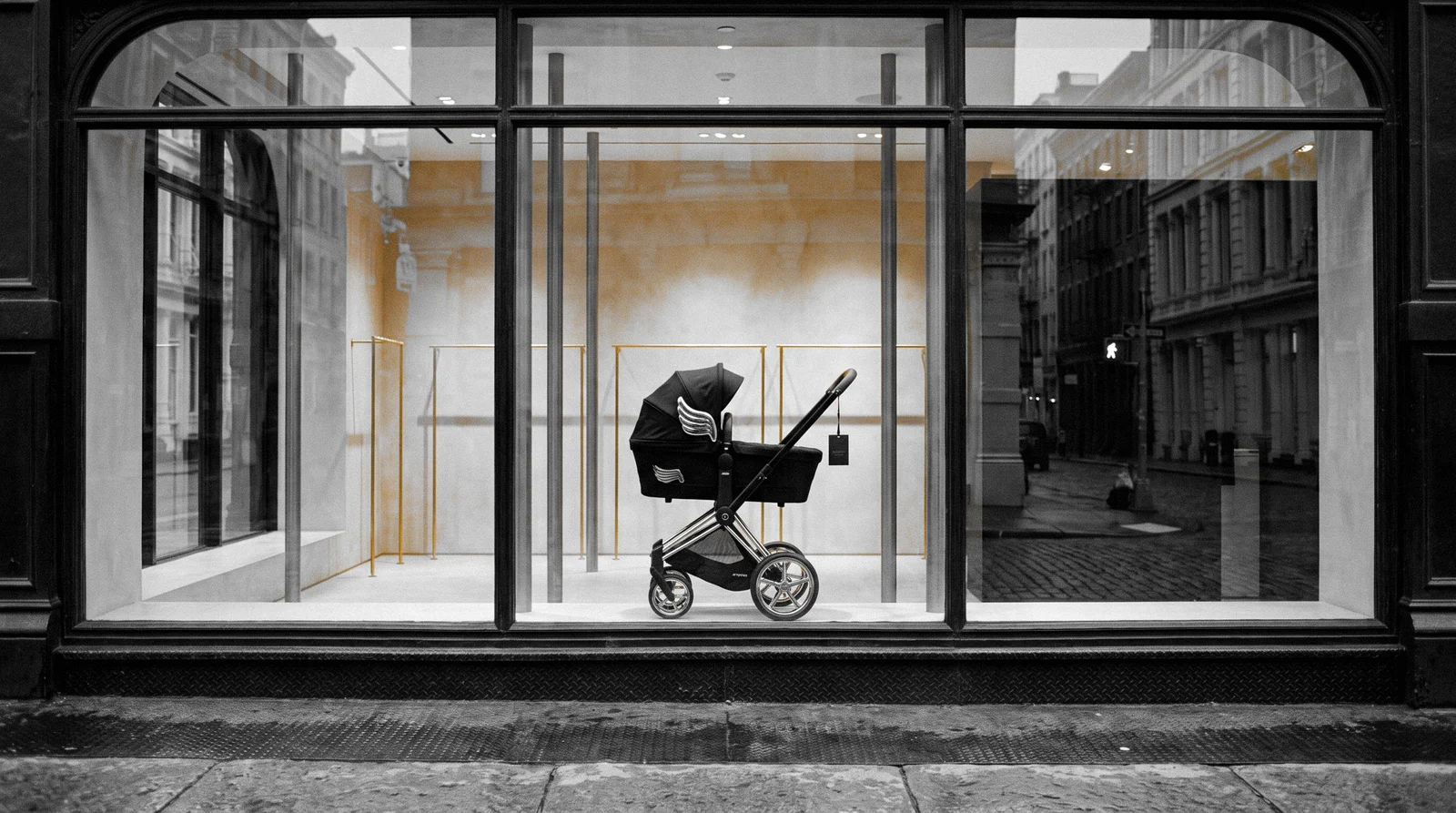 A store window on a cast-iron street in black and white, a black stroller with gold wings in the window and only the gold in colour
