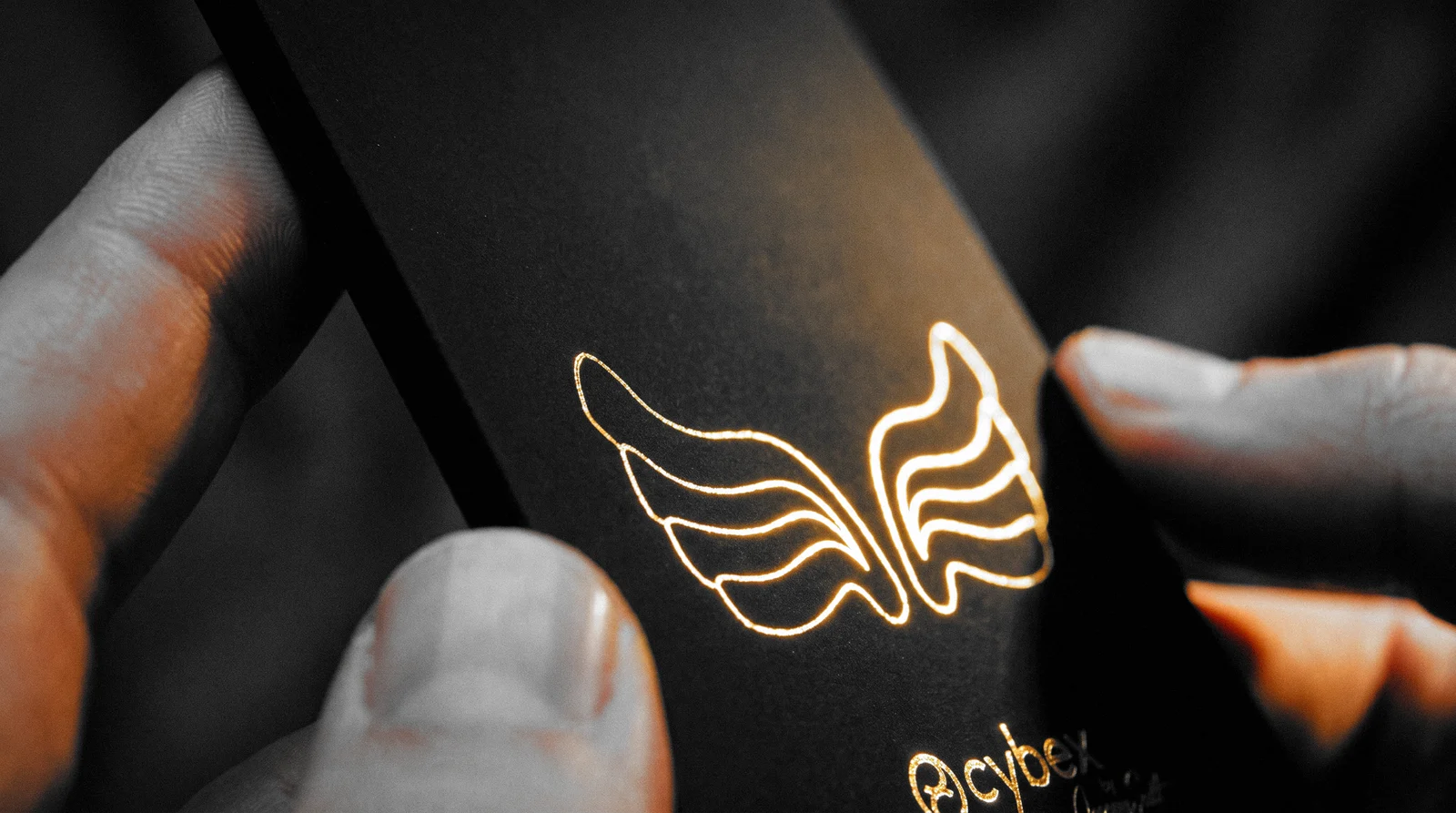 Macro of the thick black hang tag held between finger and thumb, gold foil wings catching the light on the soft-touch surface
