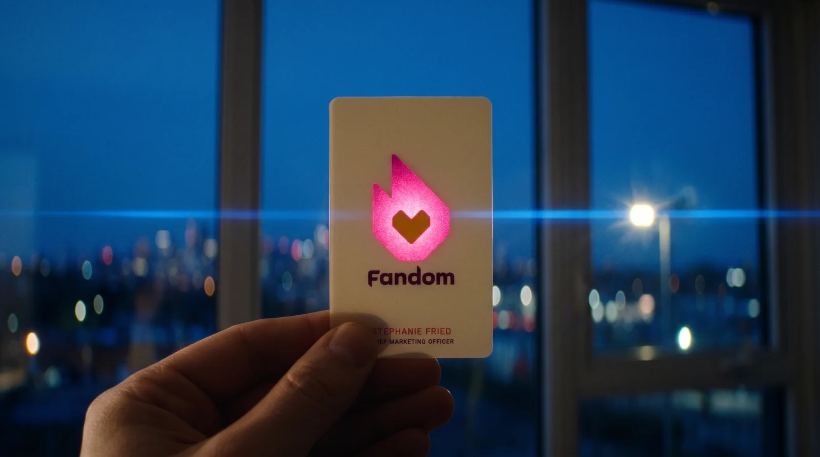 A hand holding the Fandom card against a window at blue hour with city lights behind; the flame glows