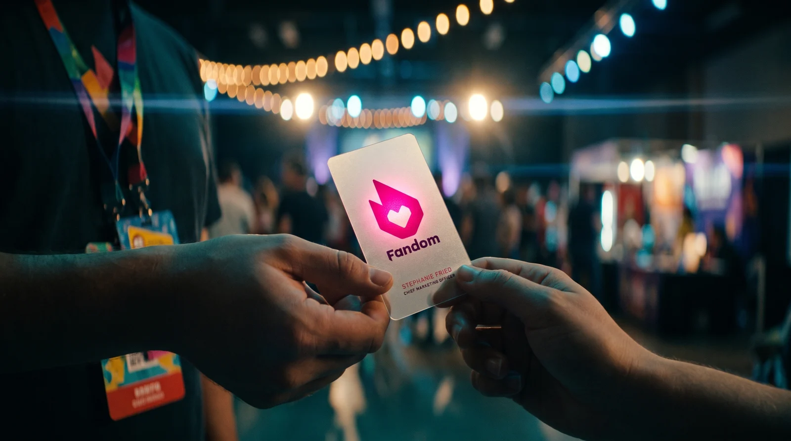 Two hands passing the Fandom card on a fan convention floor at night, lights and badges blurred behind