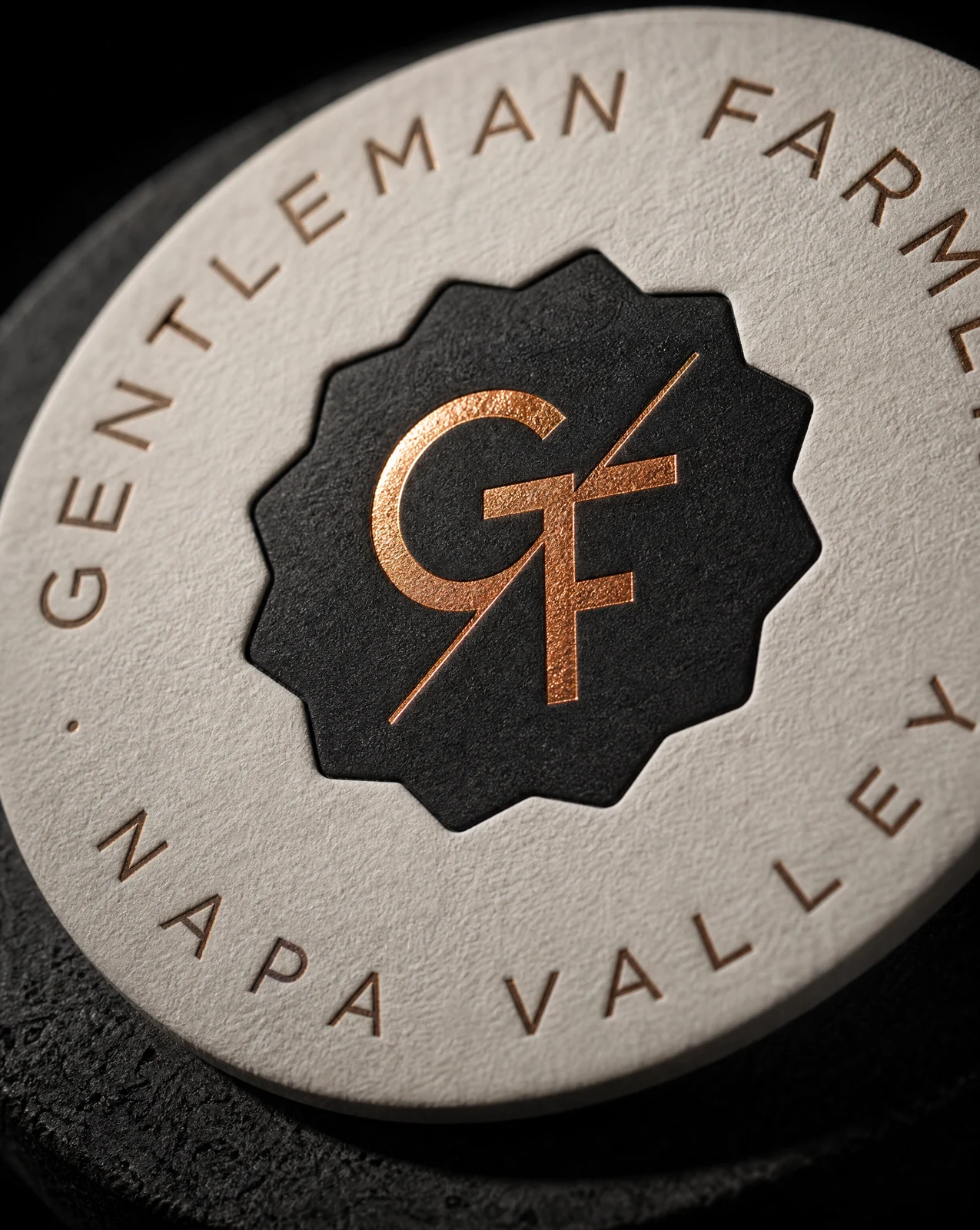 Macro of the Gentleman Farmer badge on the coaster — copper foil monogram inside a deep black letterpress seal on Gmund Heidi
