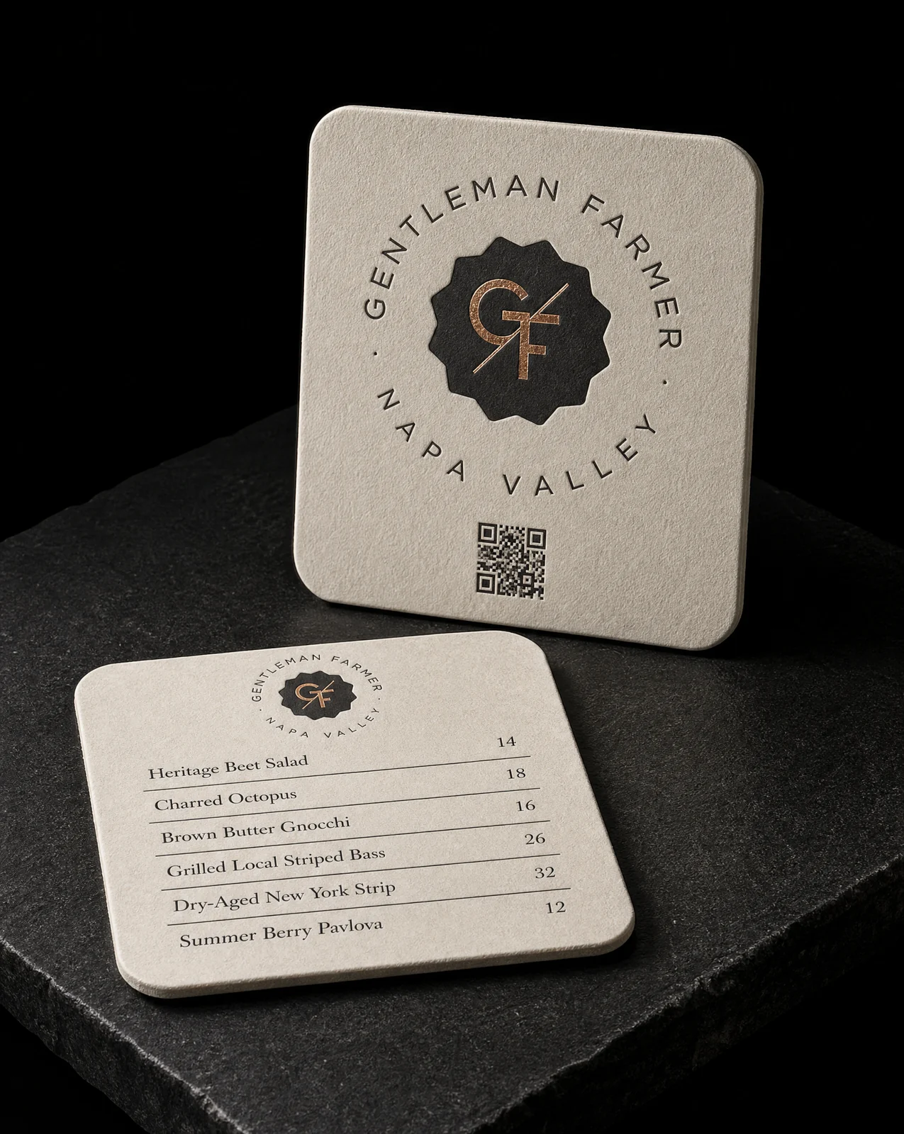 Two Gentleman Farmer coasters on warm bone — 530gsm Gmund Heidi, black letterpress badge and a copper foil monogram, rounded corners, one standing and one lying flat.