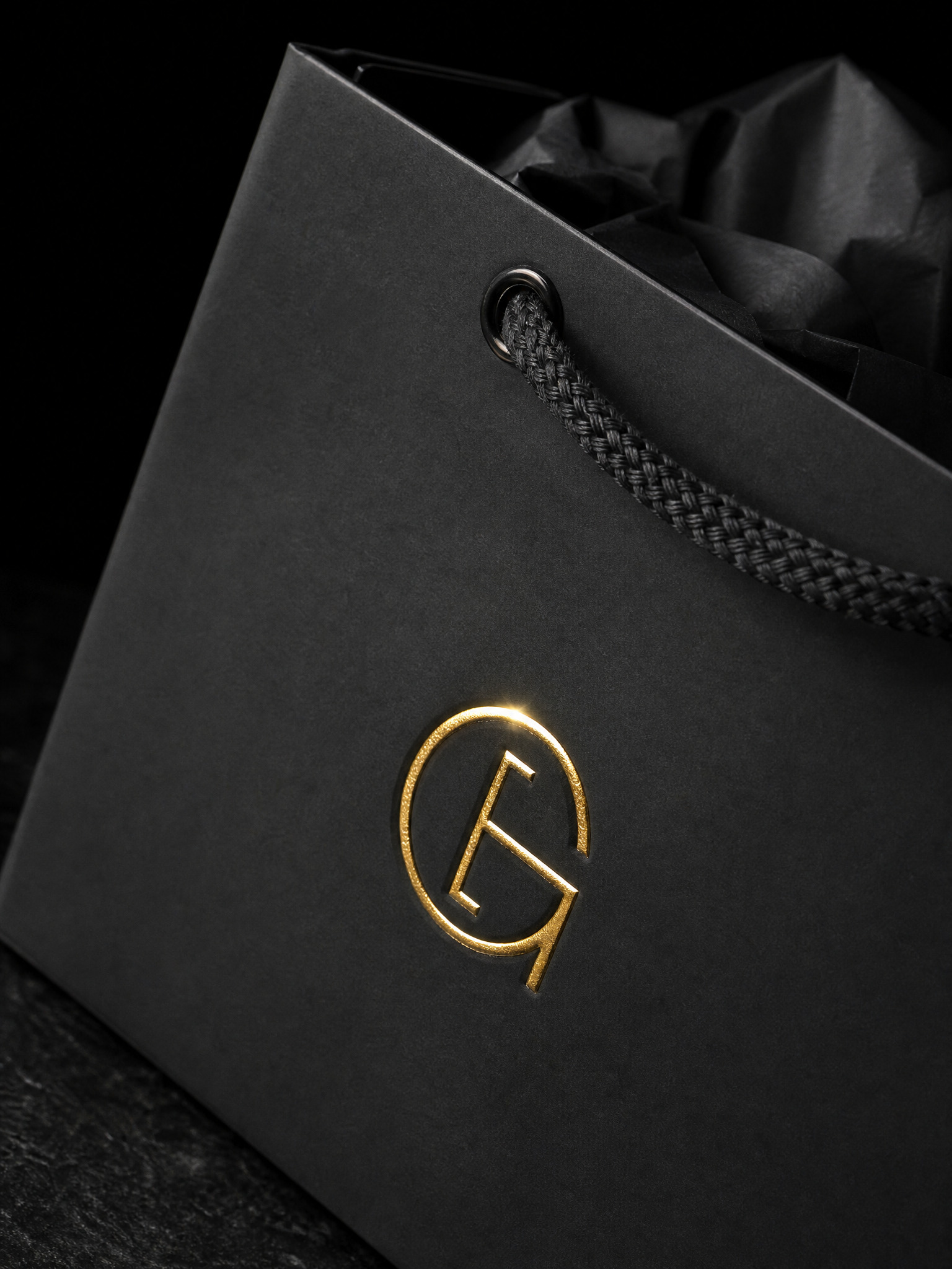 Macro: the gold foil monogram on the matte black laminate, the rope handle grommet