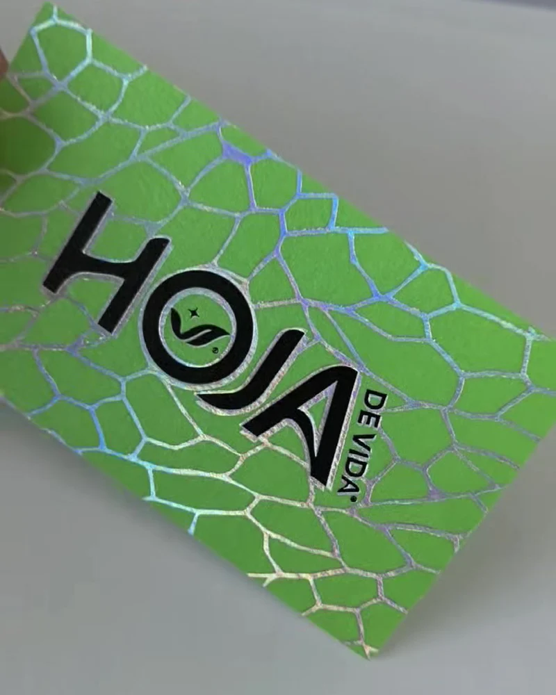 Phone photo of the real printed HOJA card: lime-green cotton with silver-rainbow holographic foil veins and a black foil HOJA DE VIDA logo