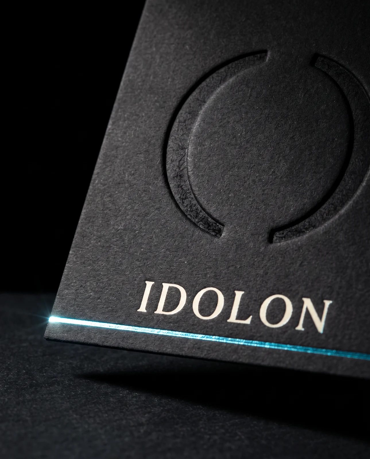 Macro of the blind deboss on dyed-through black stock, with the metallic cyan foil rule below it