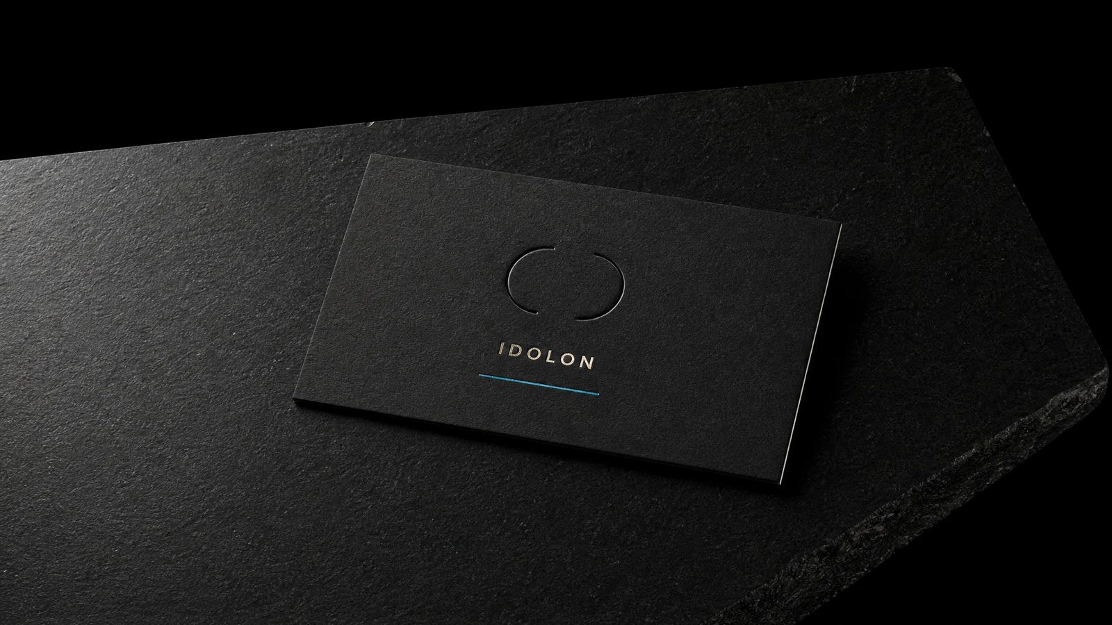 The finished IDOLON card lying on a near-black surface, the blind deboss and the cyan rule catching a single raking light