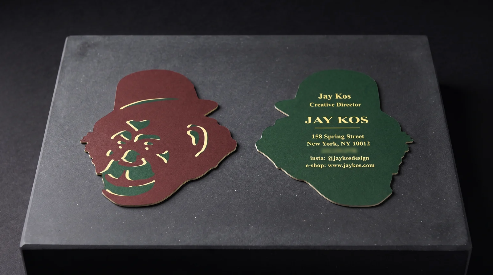 The Jay Kos die-cut card, front and back, lying on a black stone plinth: the brown monkey head in a bowler hat beside the bottle-green back with yellow type