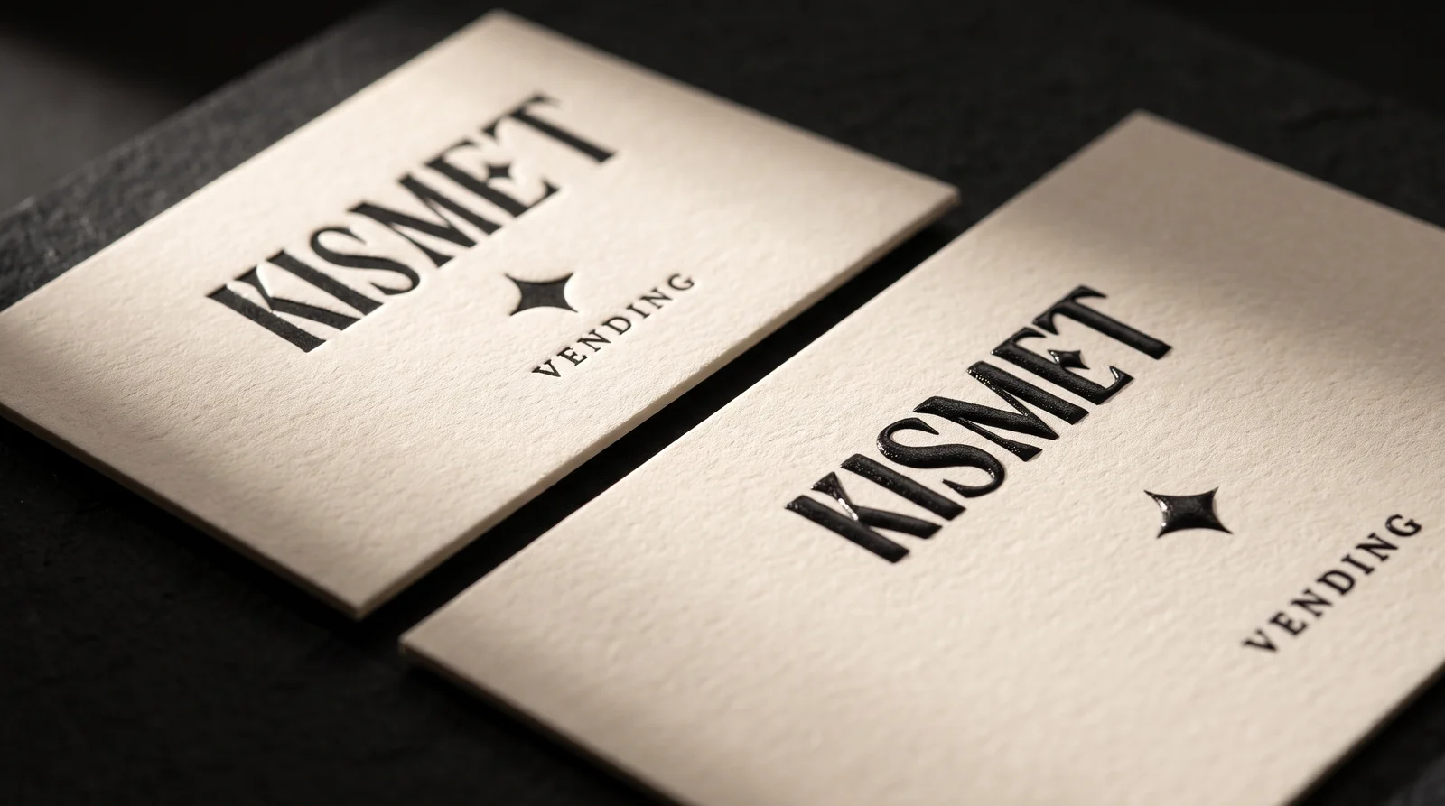 Two Kismet cards side by side under one raking light: left, the KISMET wordmark letterpressed into cotton; right, the same wordmark in raised black thermography ink. Production simulation.