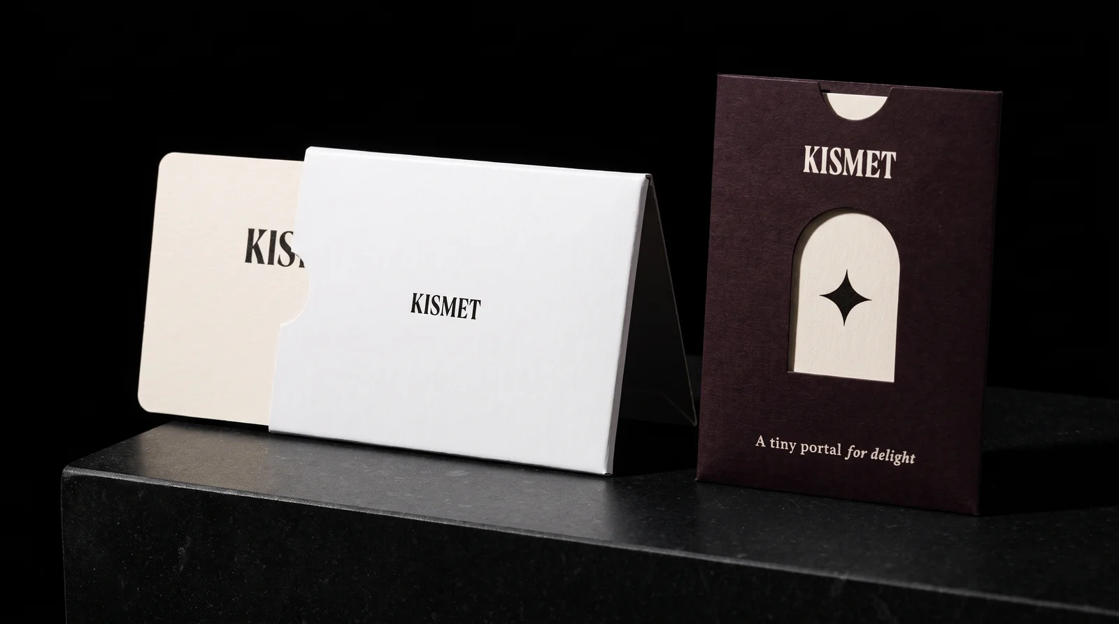 Two Kismet sleeve constructions side by side on a black plinth: a white folded sleeve printed KISMET with an off-white card half slid out, and an oxblood-black die-cut envelope sleeve with an arch window.