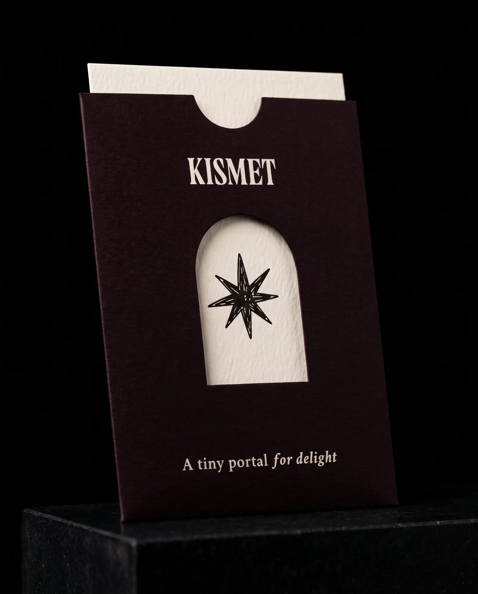 A Kismet Vending oxblood-black sleeve with a thumb notch, KISMET in off-white, an arch-shaped die-cut window framing the black eight-point star on the off-white card inside, and the line A tiny portal for delight, on a black stone plinth.