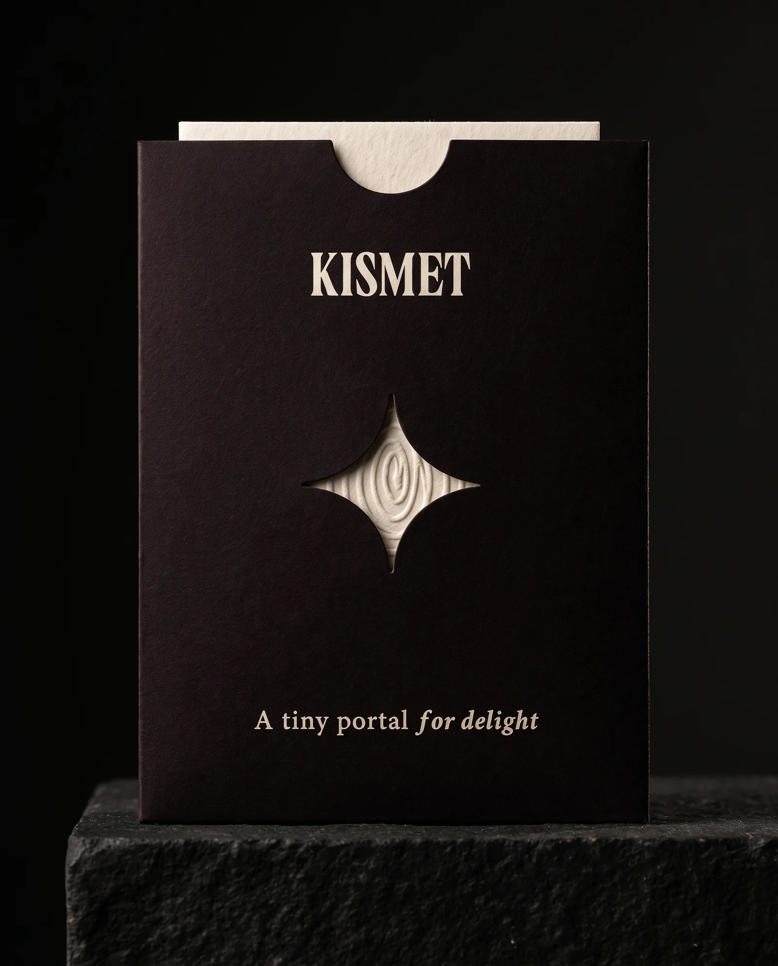 An oxblood-black Kismet sleeve with a four-point star die-cut window framing the cotton card inside, on a black plinth. Production simulation.