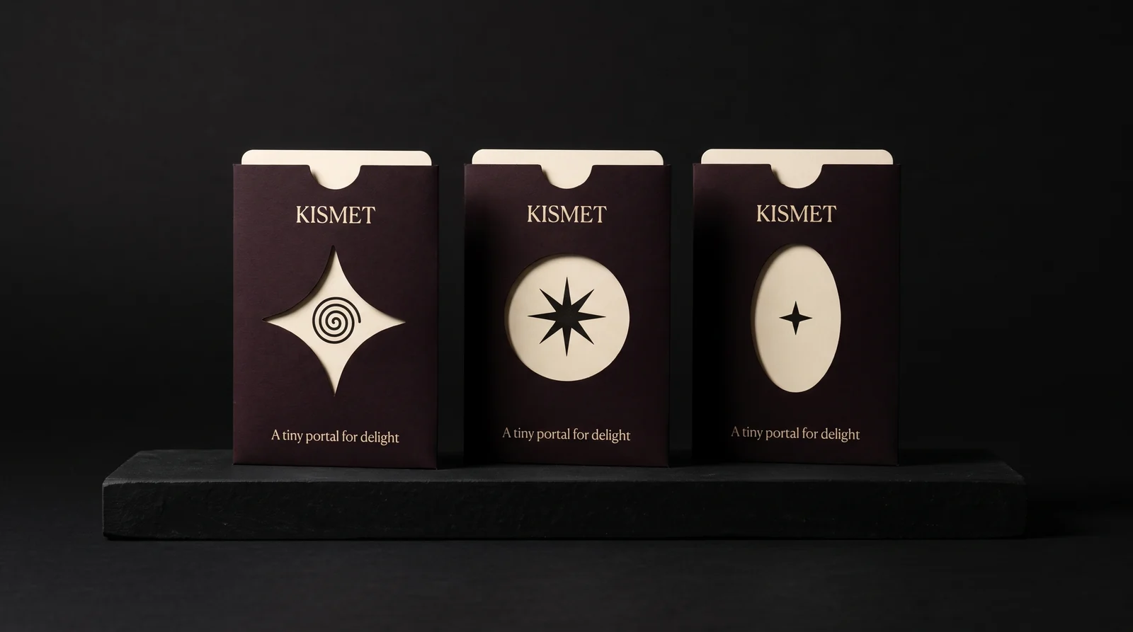 Three oxblood-black Kismet sleeves standing on a black plinth with star, circle and oval die-cut windows, each showing the cotton card inside. Production simulation.