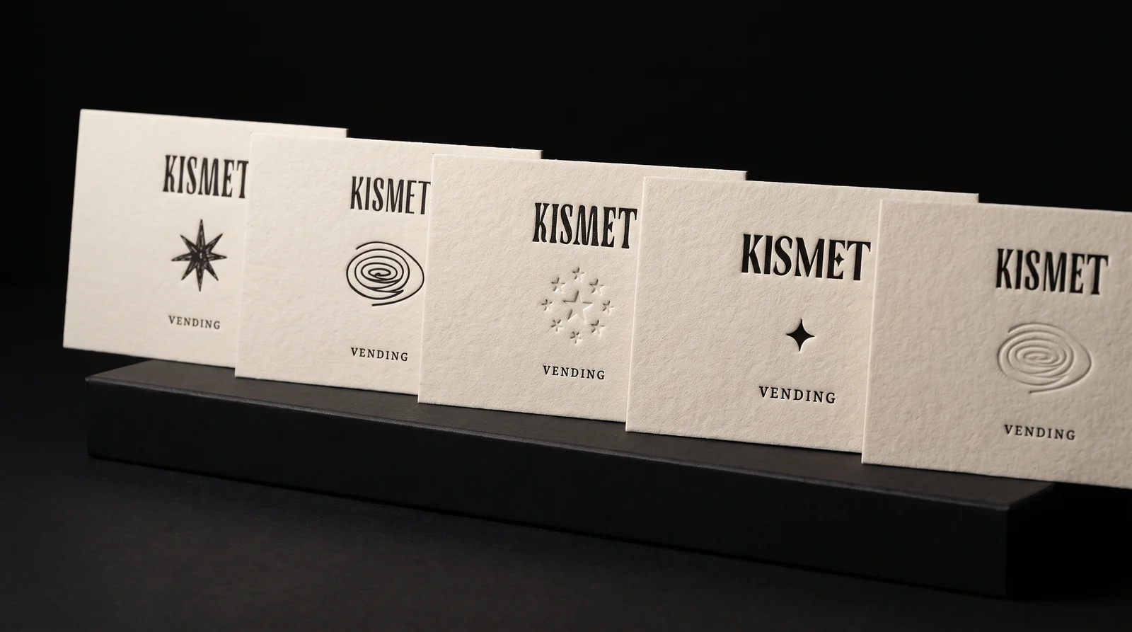 Five Kismet Vending card fronts in a row on a black plinth, each with the KISMET wordmark and a different mark: an eight-point star, a spiral, a blind debossed star cluster, a four-point star and a blind debossed spiral. Production simulation.