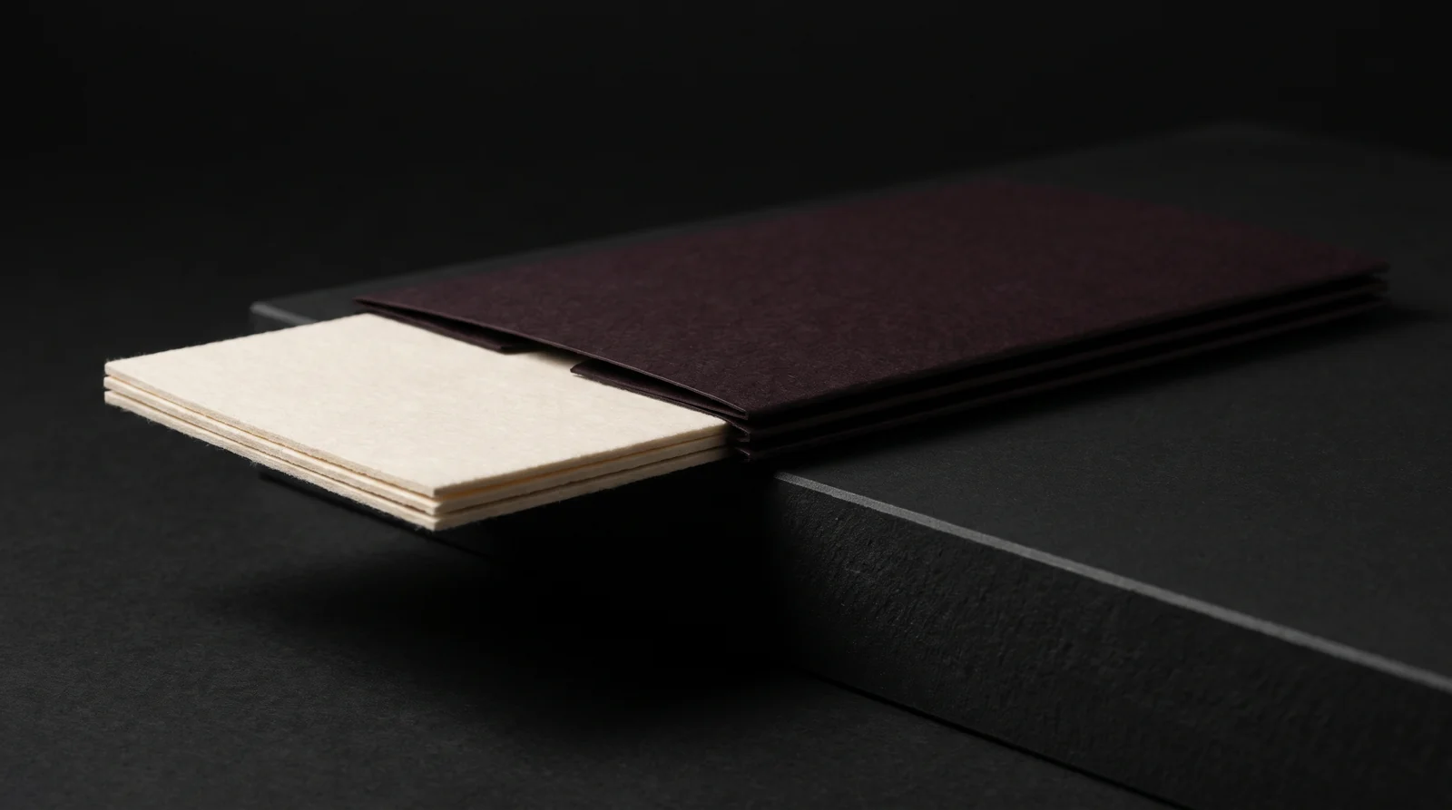 A Kismet cotton card half slid out of an oxblood-black sleeve, seen edge-on at plinth level to show the layers of paper. Production simulation.