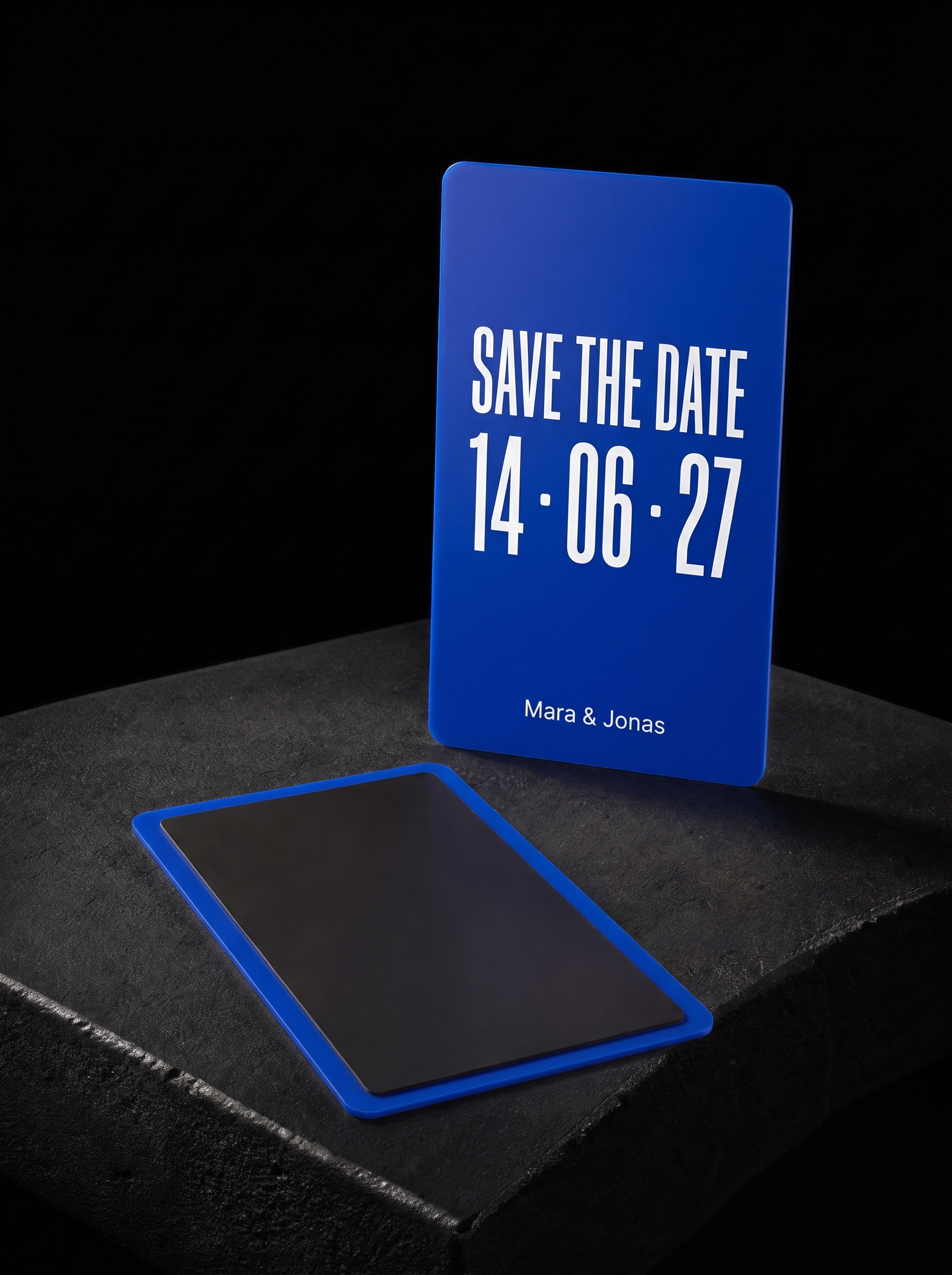 A Klein blue PVC save-the-date printed in white, standing on a slate plinth, with a second card face down showing its black magnetic backing.