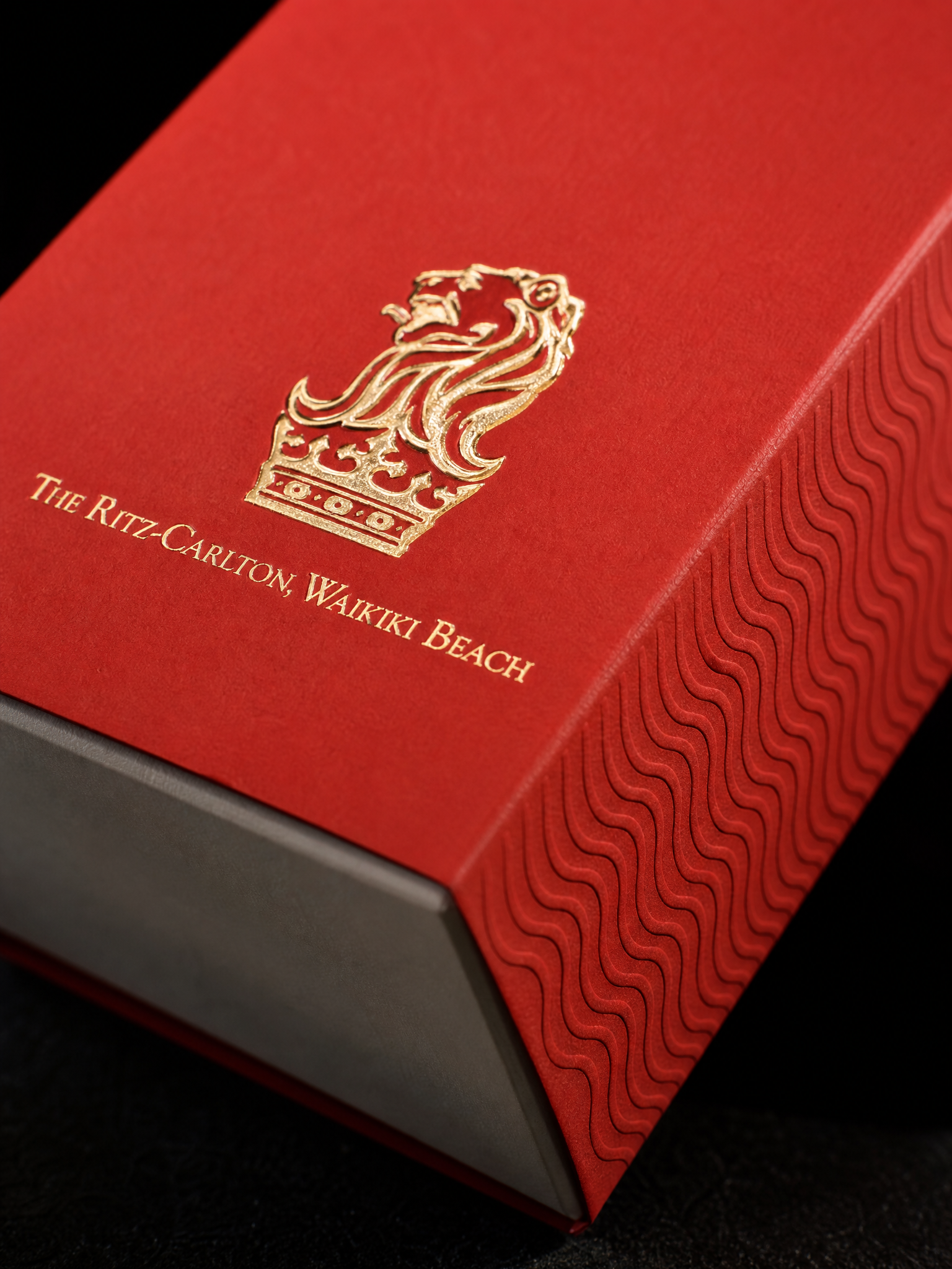 Macro: the gold foil crest on the vermilion band where it wraps the box edge, the blind-embossed wave on the side panel