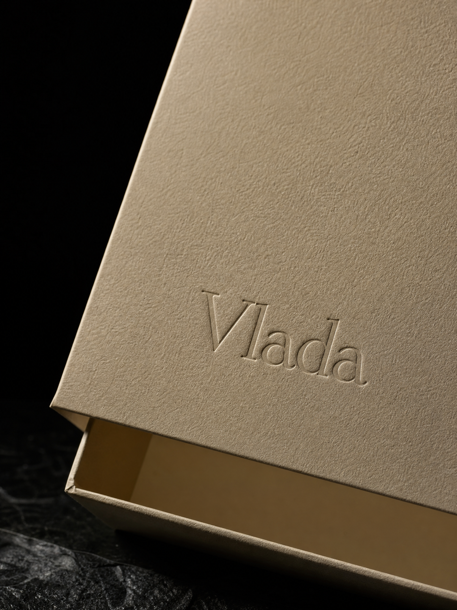 Macro: the blind-embossed VLADA rising from the sand wrap, paper fibre in raking light