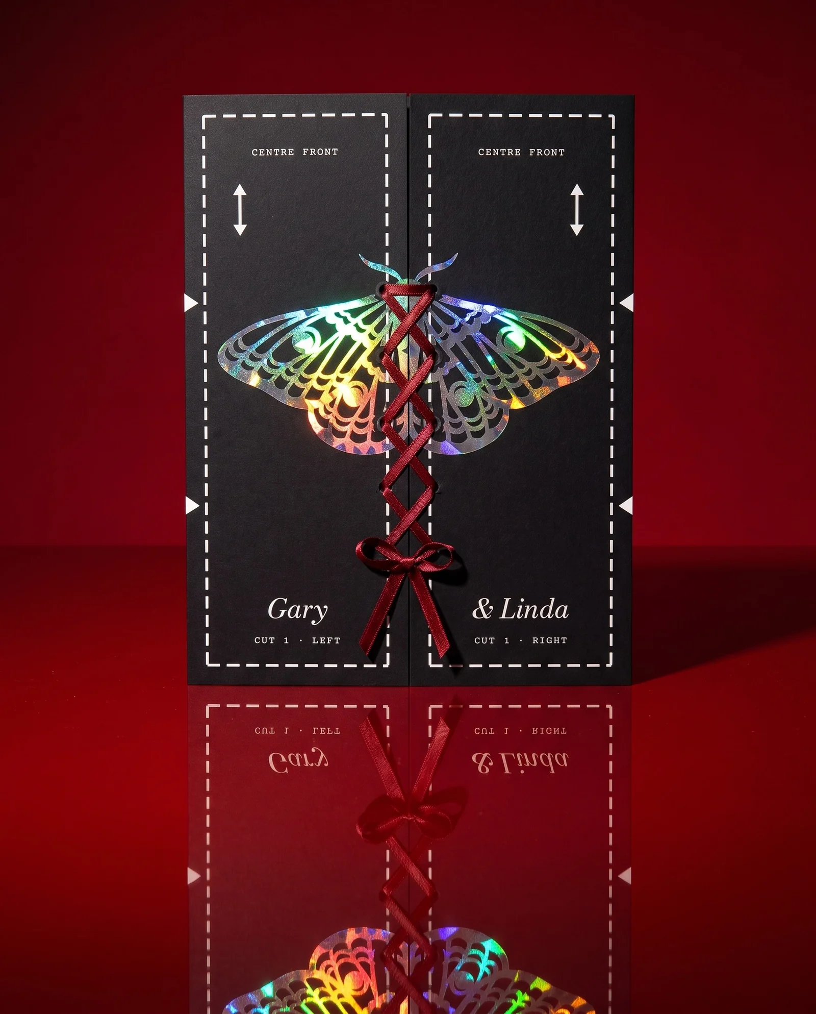 The closed black invitation laced with oxblood ribbon, standing on red lacquer with its reflection