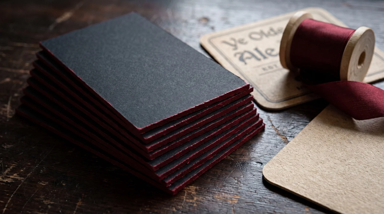 A stack of black invitations with oxblood painted edges and a spool of oxblood silk ribbon
