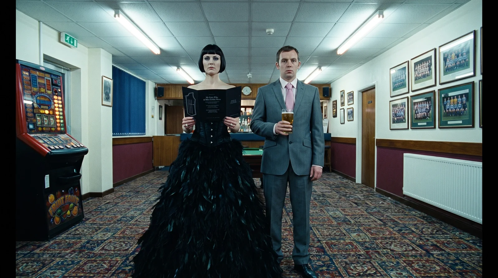The bride in a black feathered gown and the groom in a grey suit with a pint, standing in a rugby club bar