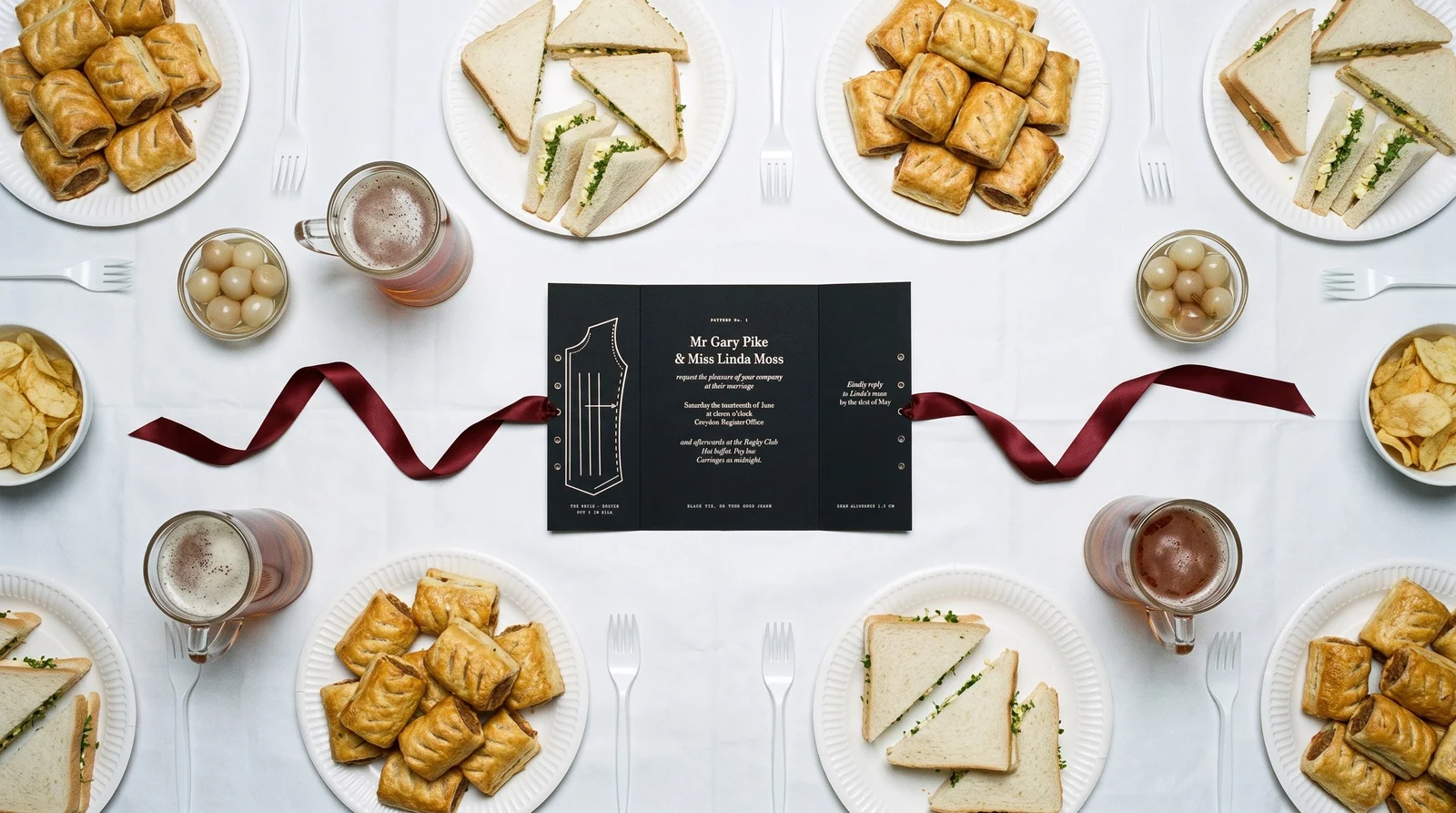 Overhead: the open black invitation on a buffet table of sausage rolls, sandwiches, pickled onions and pints