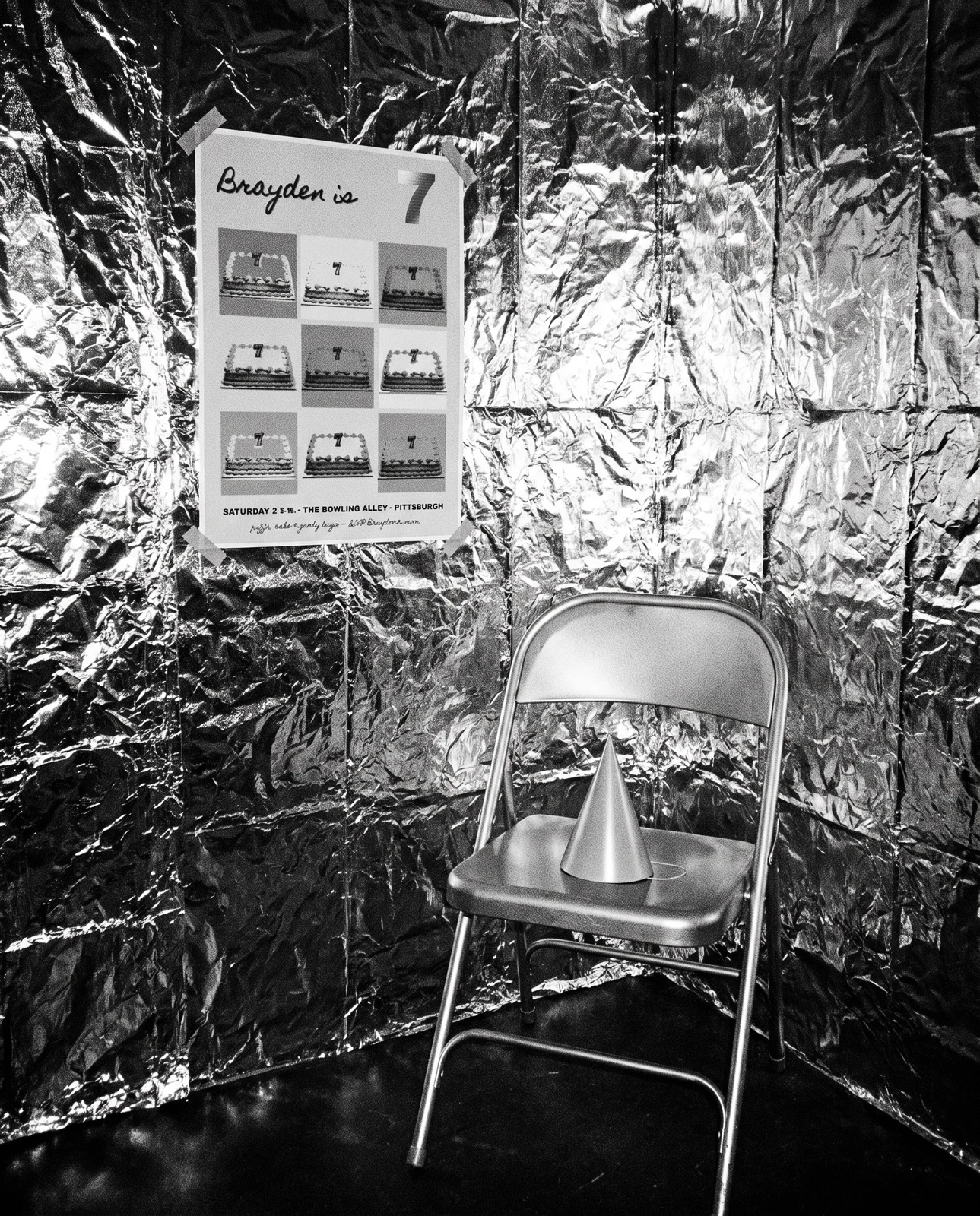 Black-and-white flash photograph of the poster taped to a foil wall, a silver chair with a party hat on it
