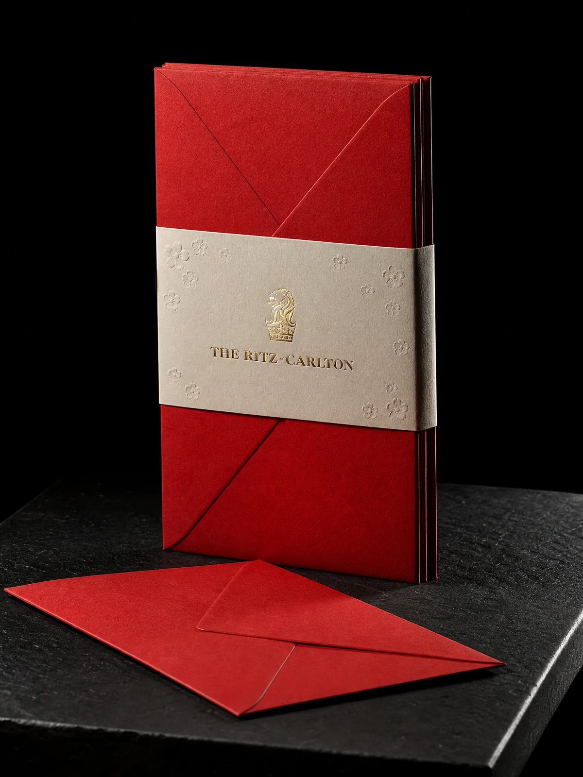 Red presentation envelopes held by a cream and gold Ritz-Carlton belly band on a black studio plinth