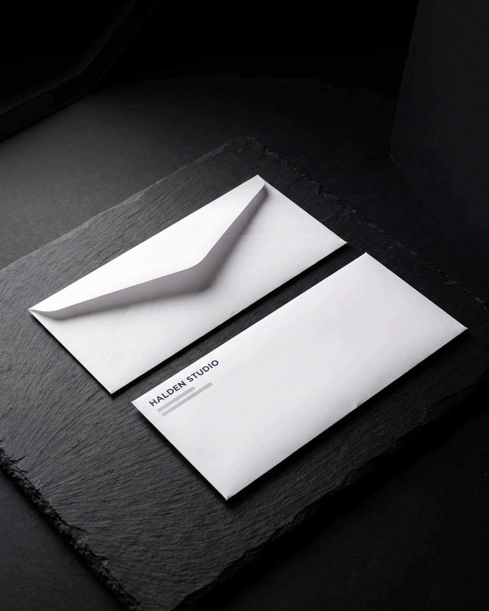 Two white #10 business envelopes on black slate, one face up with a navy "HALDEN STUDIO" return address, one showing its open flap