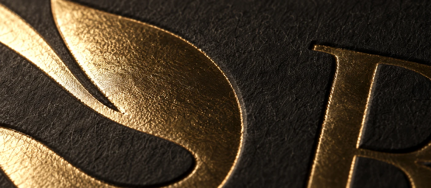 Gold hot foil stamped into black cotton card, raking light catching the mirror-bright metal and the debossed impression around it.