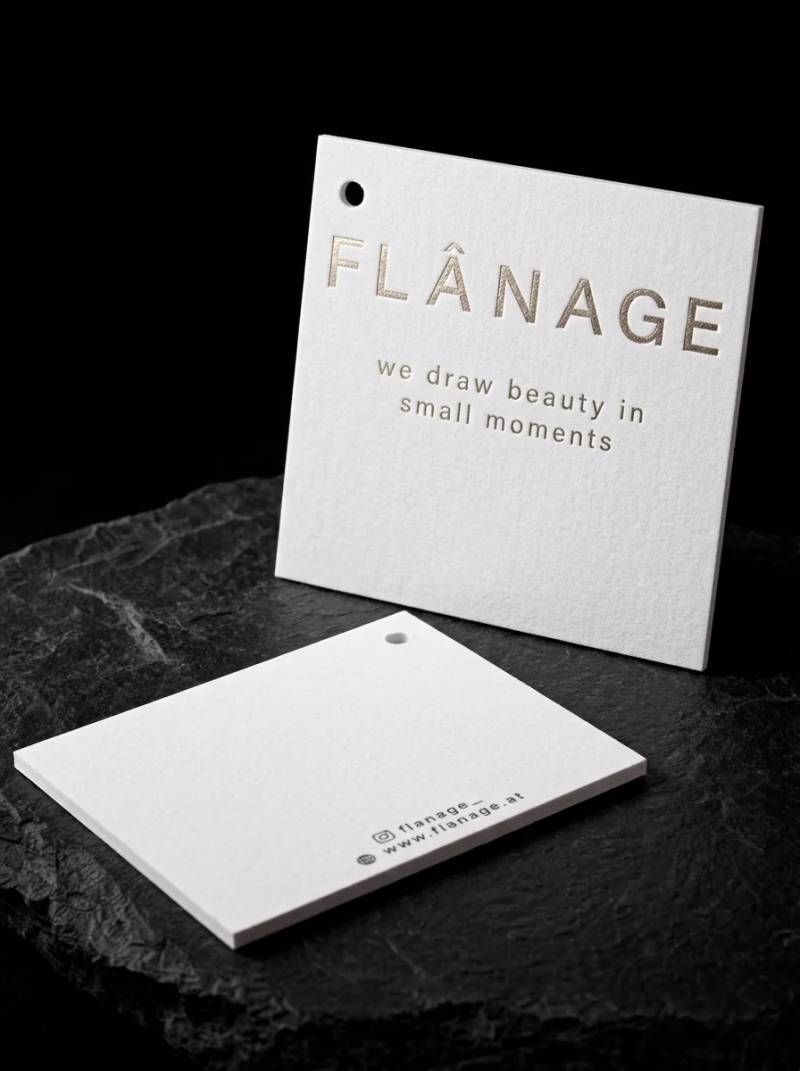 Square bright white cotton tag with a champagne foil mark, drilled for hanging.